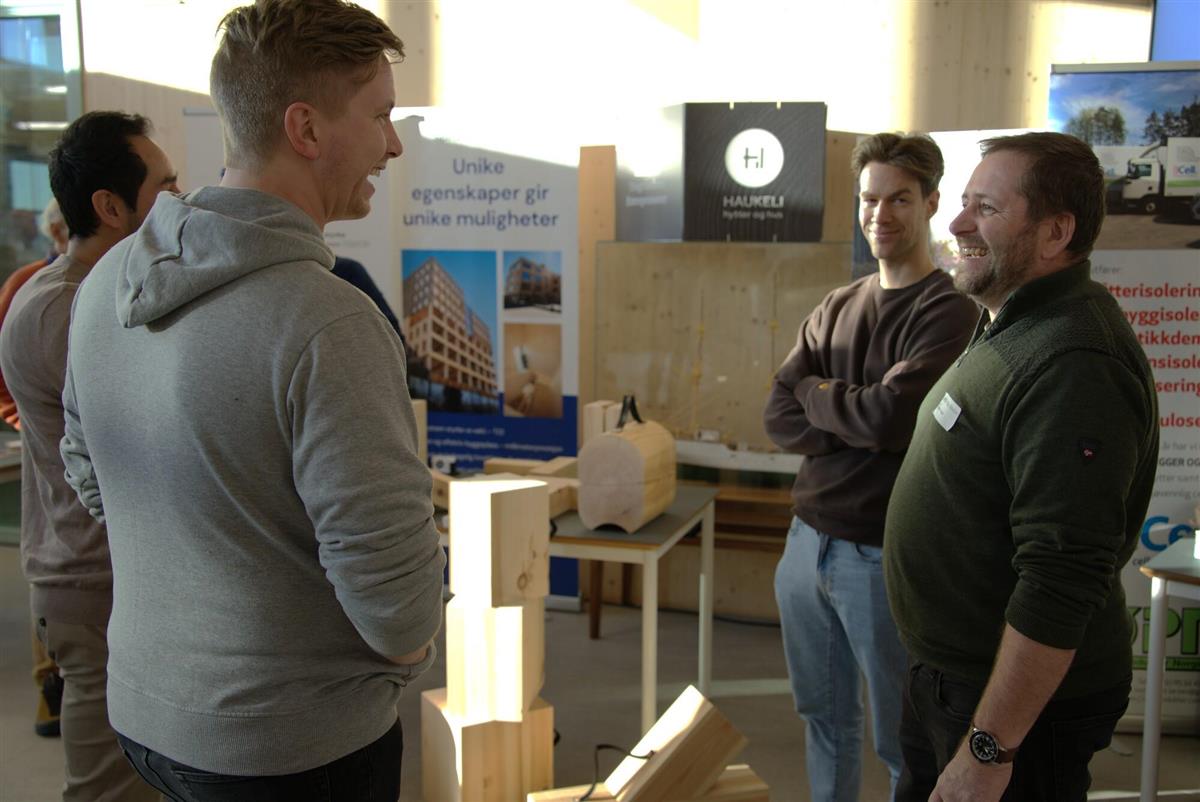 Bildet viser 2 menn som har en engasjert samtale foran en stand med trematerialer 
